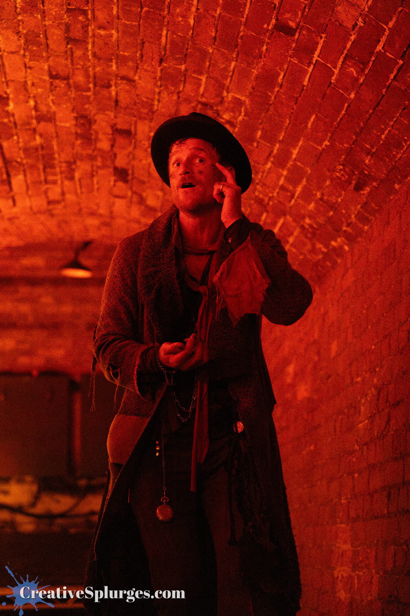 A man in period costume, wearing a wide-brimmed hat and layered clothing, stands in a dimly lit brick tunnel with red lighting. He gestures thoughtfully, possibly in conversation, as he holds a small object in his hand.
