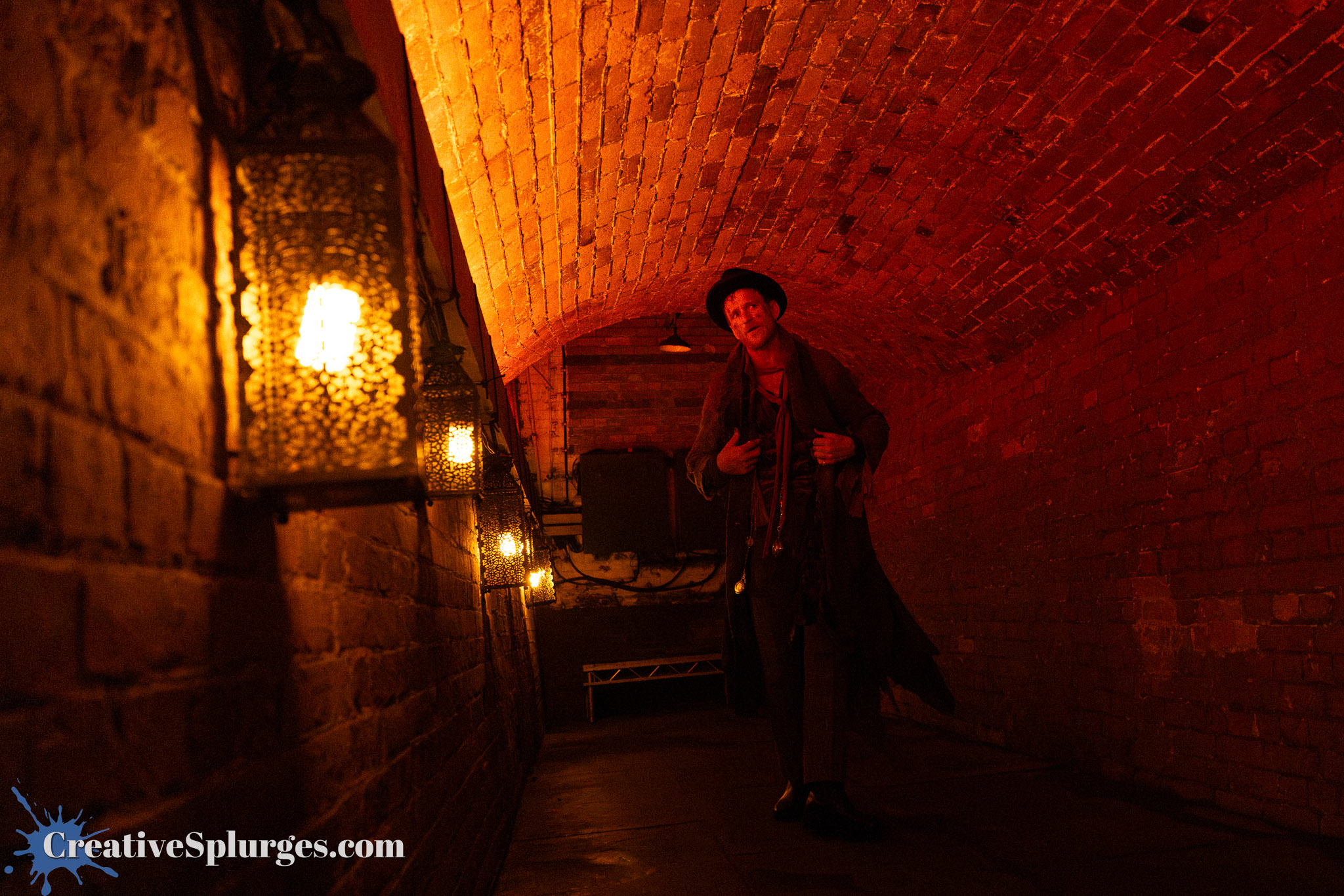 A dimly lit underground tunnel with exposed brick walls and decorative lanterns hanging along the sides. A figure dressed in a vintage costume is standing in the tunnel, creating an atmospheric and mysterious scene.