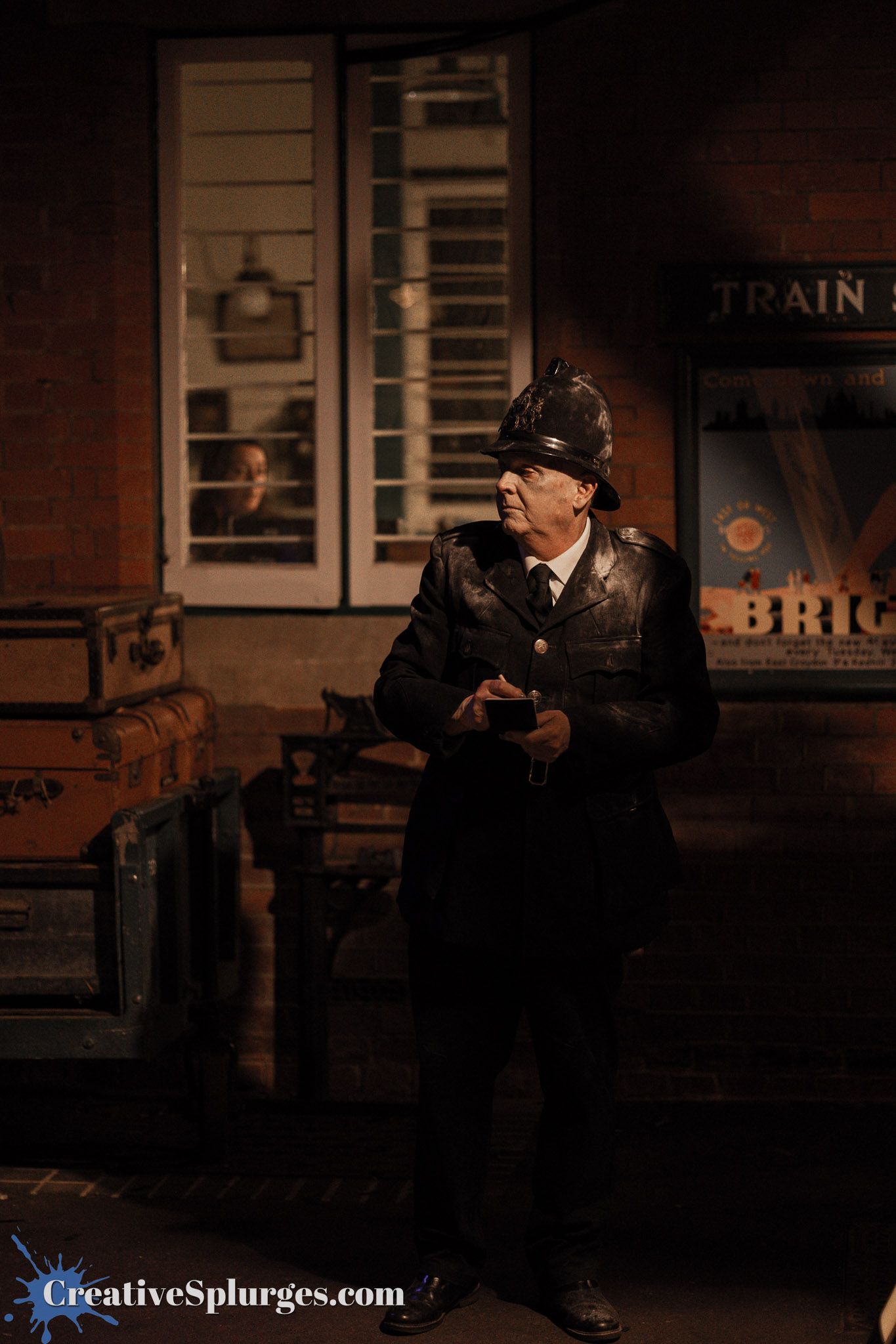 A vintage police officer standing near luggage at a train station, with a brick wall and window in the background.